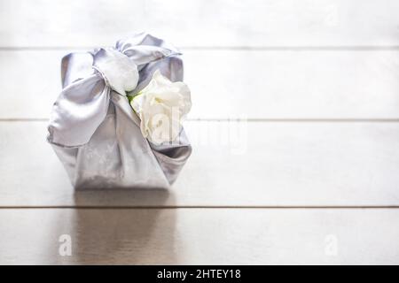 Confezione regalo trendy avvolta in tessuto di seta con tecnica Furoshiki, Eustoma fiore bianco o Lisianthus su sfondo di legno chiaro con spazio copia. Zero era Foto Stock