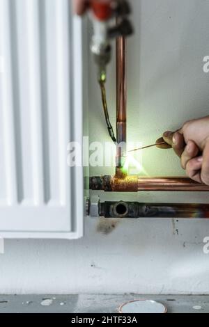 Primo piano sulle mani idraulico sconosciuto lavoratore industriale con centrale riscaldamento di tubi in rame saldatura con torcia a gas o torcia a soffiaggio accesa il muro in casa buildi Foto Stock