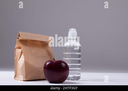 Primo piano della borsa da pranzo di carta e mela con bottiglia d'acqua sul tavolo su sfondo grigio Foto Stock