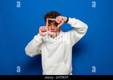 Giovane bruna bella uomo con capelli ricci indossando una felpa bianca su sfondo blu sorridente facendo cornice con mani e dita con viso felice Foto Stock