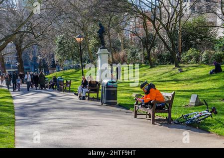 I London Memorial Gardens sono occupati di persone che camminano e si siedono su panchine lungo il percorso principale attraverso il parco. Foto Stock