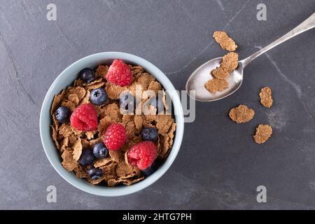 Direttamente sopra la ciotola di mirtilli e lamponi con fiocchi di mais in ciotola con un cucchiaio sull'ardesia del cibo Foto Stock