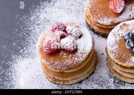 Primo piano ad angolo alto di zucchero in polvere su pancake impilati con vari frutti di bosco su ardesia alimentare Foto Stock