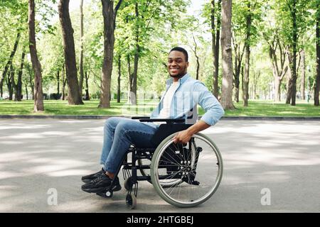 Felice giovane uomo nero in sedia a rotelle che passa il tempo al parco della città, sorridendo a macchina fotografica, a tutta lunghezza Foto Stock