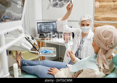Ritratto di giovane donna che guarda nello specchio e sorride con denti bianchi mentre si siede in sedia dentale alla clinica Foto Stock