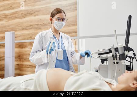 Ritratto di medico femminile utilizzando la macchina a ultrasuoni e indossare la maschera durante l'esame della donna in gravidanza in clinica Foto Stock