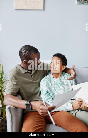 Ritratto verticale di sorridente ragazza afroamericana che fa i compiti con il padre mentre si siede sul divano Foto Stock