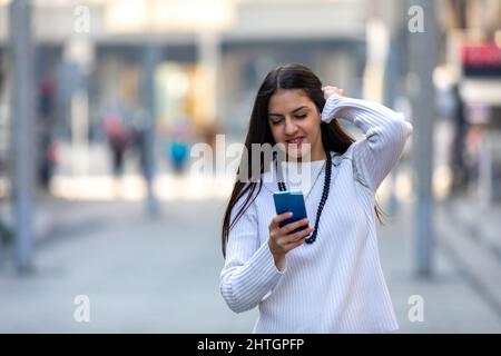 Ragazza sorridente che guarda lo smartphone, toccando e pettinando i capelli usando il cellulare come uno specchio per la strada Foto Stock