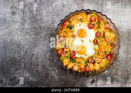 Cuocere le patate al forno con salsicce, formaggio e uova in una teglia di vetro su fondo grigio scuro. Vista dall'alto, disposizione piatta Foto Stock