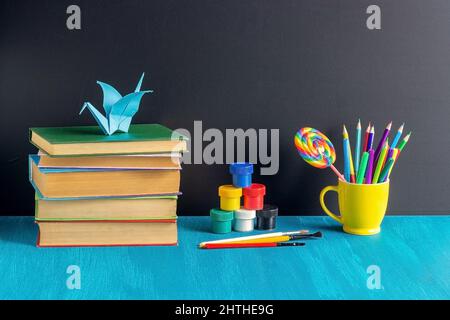 Posto di lavoro degli allievi. Libri cartoleria vernice gouache e origami gru su tavola blu su sfondo nero lavagna concetto Educazione e ritorno a scuola. Foto Stock