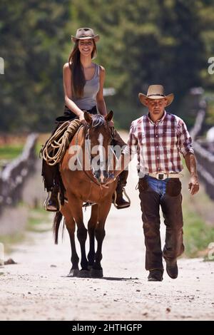 Equitazione 101. Una giovane cowgirl attraente che guida un cavallo su un ranch mentre un cowboy cammina accanto. Foto Stock