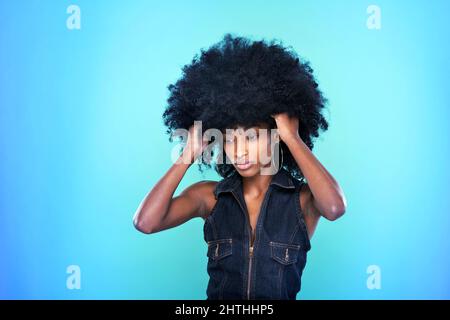 Elegante donna afroamericana in abito in denim che mantiene le mani in capelli ricci su sfondo blu in Vivid studio Foto Stock