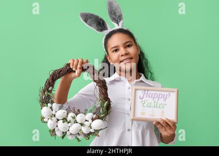 Carina ragazza afroamericana con orecchie conigliate, biglietto di auguri e corona di Pasqua su sfondo colorato Foto Stock