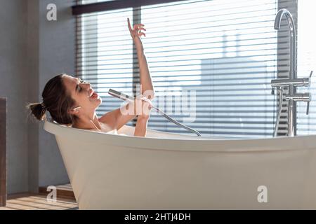 Allegra giovane donna che ascolta musica e canto mentre si rilassa nella vasca da bagno Foto Stock
