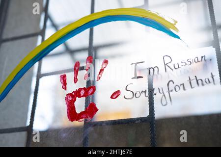 Foto sulla finestra con barre di bandiera Ucranian come un segno arcobaleno e una stampa a mano rossa con un'iscrizione di scusa sono russo. Foto Stock