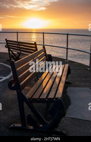 Le panchine vuote si affacciano sul mare, bagnate dal bagliore di una calda alba dorata. Buona simmetria e la luce ambra e arancione si combinano con le ombre Foto Stock