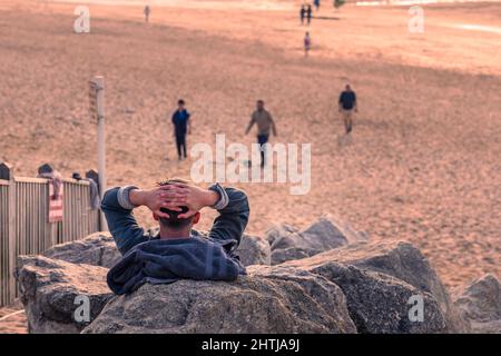 Un uomo seduto su rocce rilassandosi e guardando la luce tarda sera su Fistral a Newquay in Cornovaglia. Foto Stock