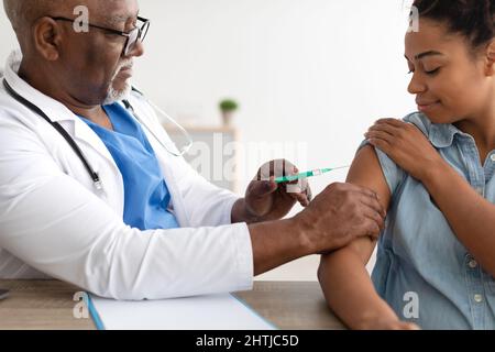 Donna nera che riceve l'iniezione di vaccino seduta con il medico indoor Foto Stock