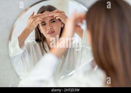 Problema Skin. Sconvolto giovane femmina schioccante pimple sulla fronte vicino a Mirror Foto Stock