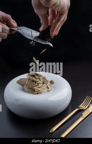 Mani di raccolto anonimo chef grattugiare deliziosi tartufi neri su pasta servita in piatto bianco vicino alle posate nel ristorante moderno Foto Stock