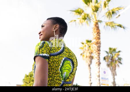 Da sotto di fiducioso felice giovane donna africana con capelli corti scuri in abito luminoso estate guardando lontano mentre in piedi vicino alle palme sulla strada o della città Foto Stock