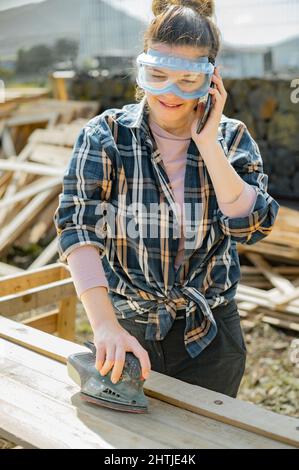 Donna in occhiali protettivi che parlano il telefono mentre carteggiava assi di legno con levigatrice orbitale in campagna il giorno d'estate Foto Stock