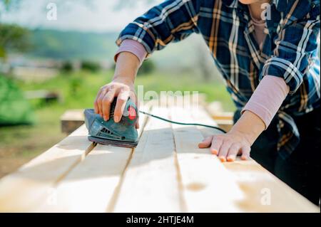 Coltivare l'artigiano femminile utilizzando una speciale levigatrice orbitale elettrica per levigare le tavole di legno mentre si lavora in campagna il giorno d'estate Foto Stock