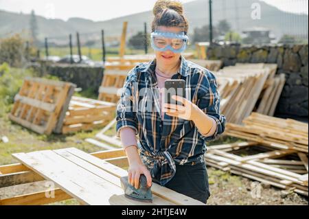 Donna in occhiali protettivi messaggi di testo sul cellulare mentre carteggiatura assi di legno con levigatrice orbitale in campagna il giorno d'estate Foto Stock