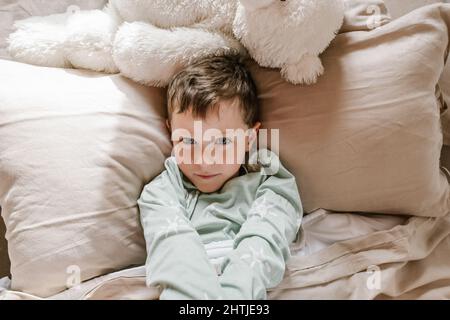 Da sopra ragazzo in pajama guardando lontano mentre si sdraia su un comodo letto in camera da letto luminosa con finestra Foto Stock