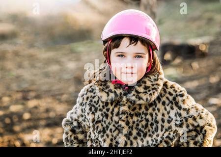 Adorabile ragazza in casco protettivo rosa e caldo cappotto in pelliccia guardando la macchina fotografica mentre si trova in campagna su sfondo sfocato Foto Stock