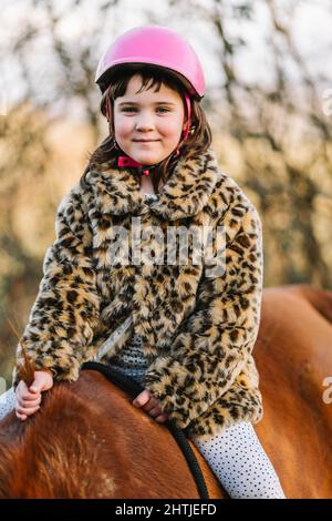 Vista laterale della ragazza positiva in casco protettivo rosa guardando la macchina fotografica mentre si guida cavallo marrone in zona rurale con alberi in campagna Foto Stock