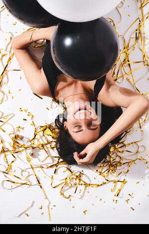 Da sopra di ragazza allegra con palloncini neri che giacciono con occhi chiusi sul pavimento con tinsel durante la festa in studio Foto Stock