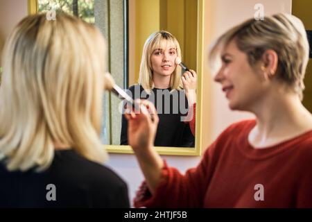 Visagista femminile positivo applicare polvere sul viso di giovane donna attraente con pennello mentre si siede in salone con specchio durante la procedura di bellezza Foto Stock