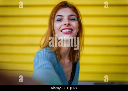 Bella sorridente giovane donna che guarda la macchina fotografica mentre si prende autoritratto sulla strada vicino al muro giallo Foto Stock