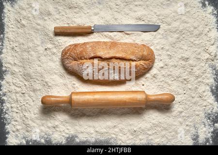 Vista dall'alto, filone a telaio pieno di pane appena sfornato con spilla in legno e coltello posto sulla farina bianca in una stanza luminosa Foto Stock