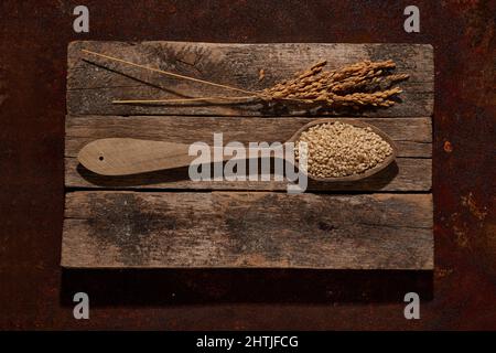 Vista dall'alto cucchiaio di legno con semi di sesamo crudi su assi di legno con orecchie di riso poste sul tavolo in camera luminosa Foto Stock