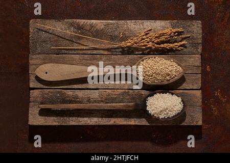 Vista dall'alto cucchiai di legno completi con set di semi di riso e sesamo con grani di orzo posti sul tagliere di cesoia vicino con spighe di riso Foto Stock