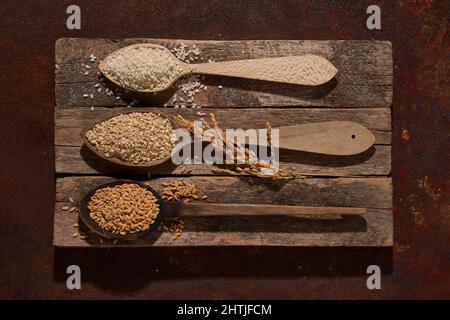 Vista dall'alto cucchiai di legno completi con set di semi di riso e sesamo con grani di orzo posti sul tagliere di cesoia vicino con spighe di riso Foto Stock