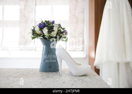 Scarpe bianche nuziali e mazzo di fiori messi sul tavolo contro sfondo sfocato di abito da sposa in camera chiara Foto Stock