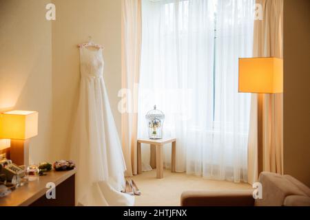 Abito da sposa bianco appeso alla parete sopra le scarpe da sposa vicino ai fiori sulla sedia e lampade incandescenti in appartamento luminoso Foto Stock