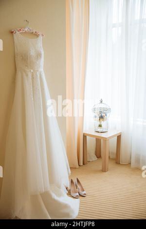 Abito da sposa bianco appeso alla parete sopra le scarpe da sposa vicino ai fiori sulla sedia e lampade incandescenti in appartamento luminoso Foto Stock