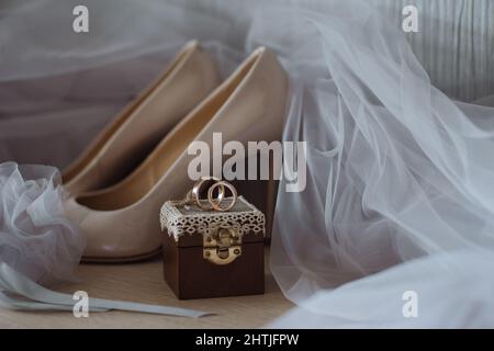 Composizione di scarpe beige con tacco alto poste contro un paio di anelli dorati su scatola decorata e velo da sposa Foto Stock
