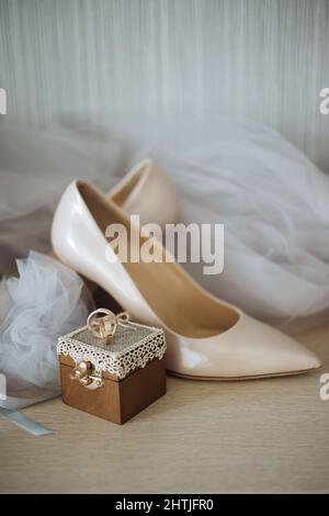 Composizione di scarpe beige con tacco alto poste contro un paio di anelli dorati su scatola decorata e velo da sposa Foto Stock