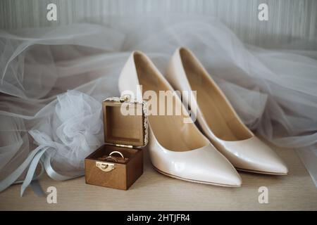 Composizione di scarpe beige con tacco alto poste contro un paio di anelli dorati su scatola decorata e velo da sposa Foto Stock