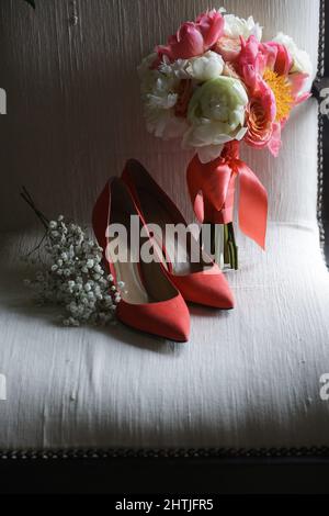 Scarpe da matrimonio rosse poste su poltrona bianca con bouquet di fiori in fiore legati con nastro in camera luminosa durante la preparazione delle vacanze Foto Stock