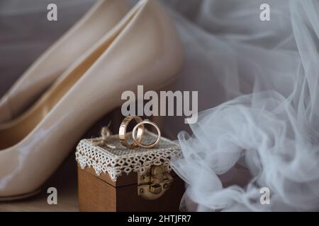 Composizione di scarpe beige con tacco alto poste contro un paio di anelli dorati su scatola decorata e velo da sposa Foto Stock