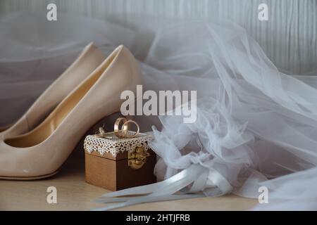 Composizione di scarpe beige con tacco alto poste contro un paio di anelli dorati su scatola decorata e velo da sposa Foto Stock