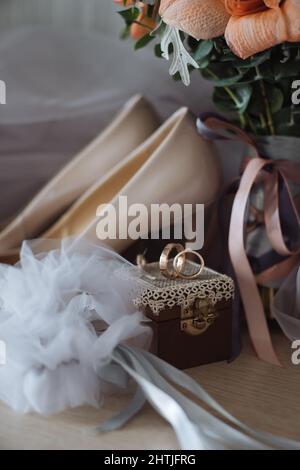 Mazzo di fiori freschi e scarpe nuziali bianche con anelli su piccola scatola decorata posta contro il velo Foto Stock