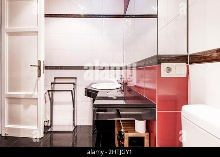 Bagno con vanity in marmo nero e lavandino in porcellana bianca con specchio in vetro quadrato senza cornice e piastrelle rosse e bianche Foto Stock