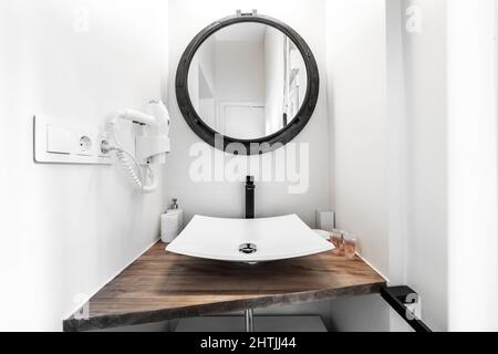 Piccolo bagno con piano in legno, lavello in porcellana senza inserto, specchio rotondo con cornice nera e asciugacapelli bianco a parete Foto Stock